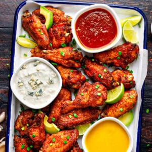 A tray of crispy chicken wings garnished with lime wedges and chopped chives, served with three dipping sauces: blue cheese, red sauce, and honey mustard. The tray sits on a dark wooden table.