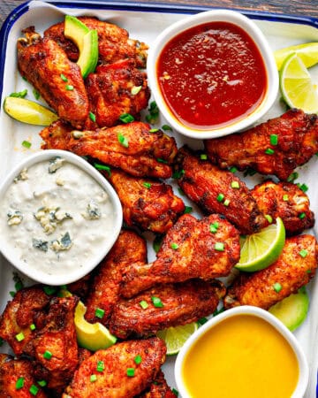 A tray of crispy chicken wings garnished with lime wedges and chopped chives, served with three dipping sauces: blue cheese, red sauce, and honey mustard. The tray sits on a dark wooden table.