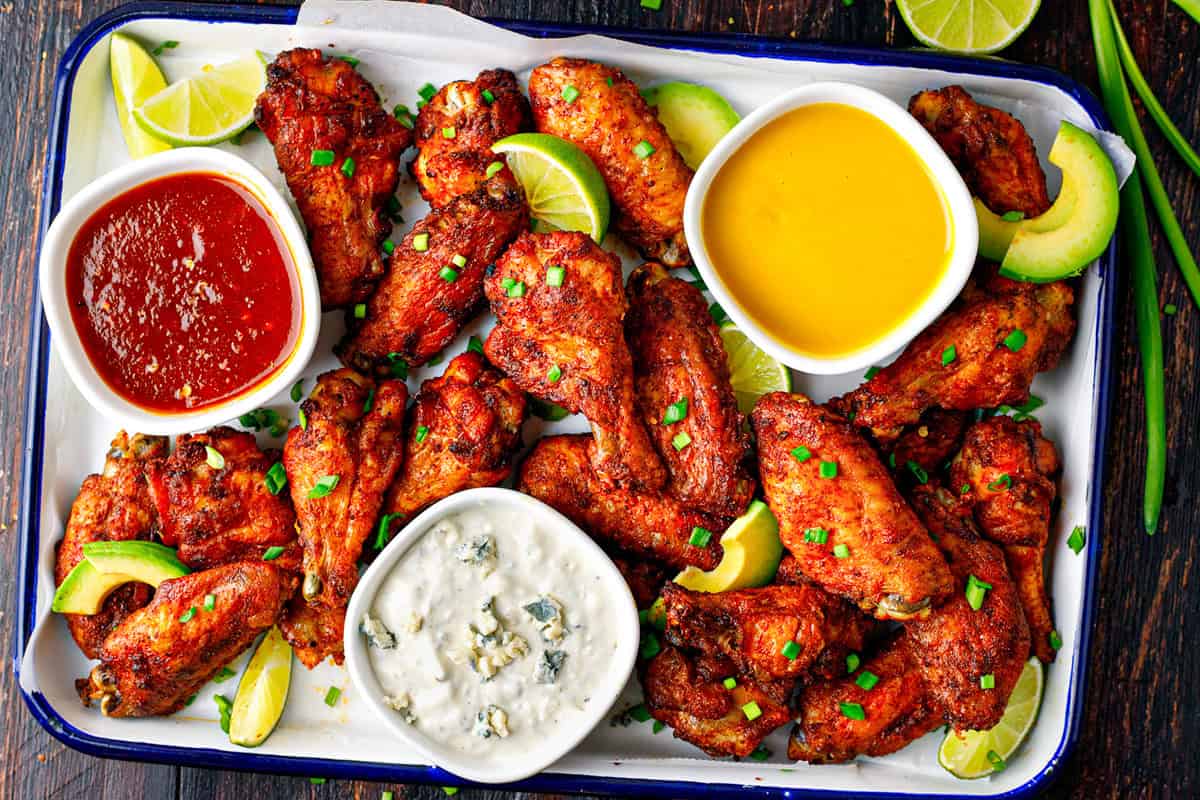 A tray of crispy chicken wings garnished with green onions and lime wedges, served with three dipping sauces: red, yellow, and creamy white with blue cheese crumbles, alongside avocado slices.