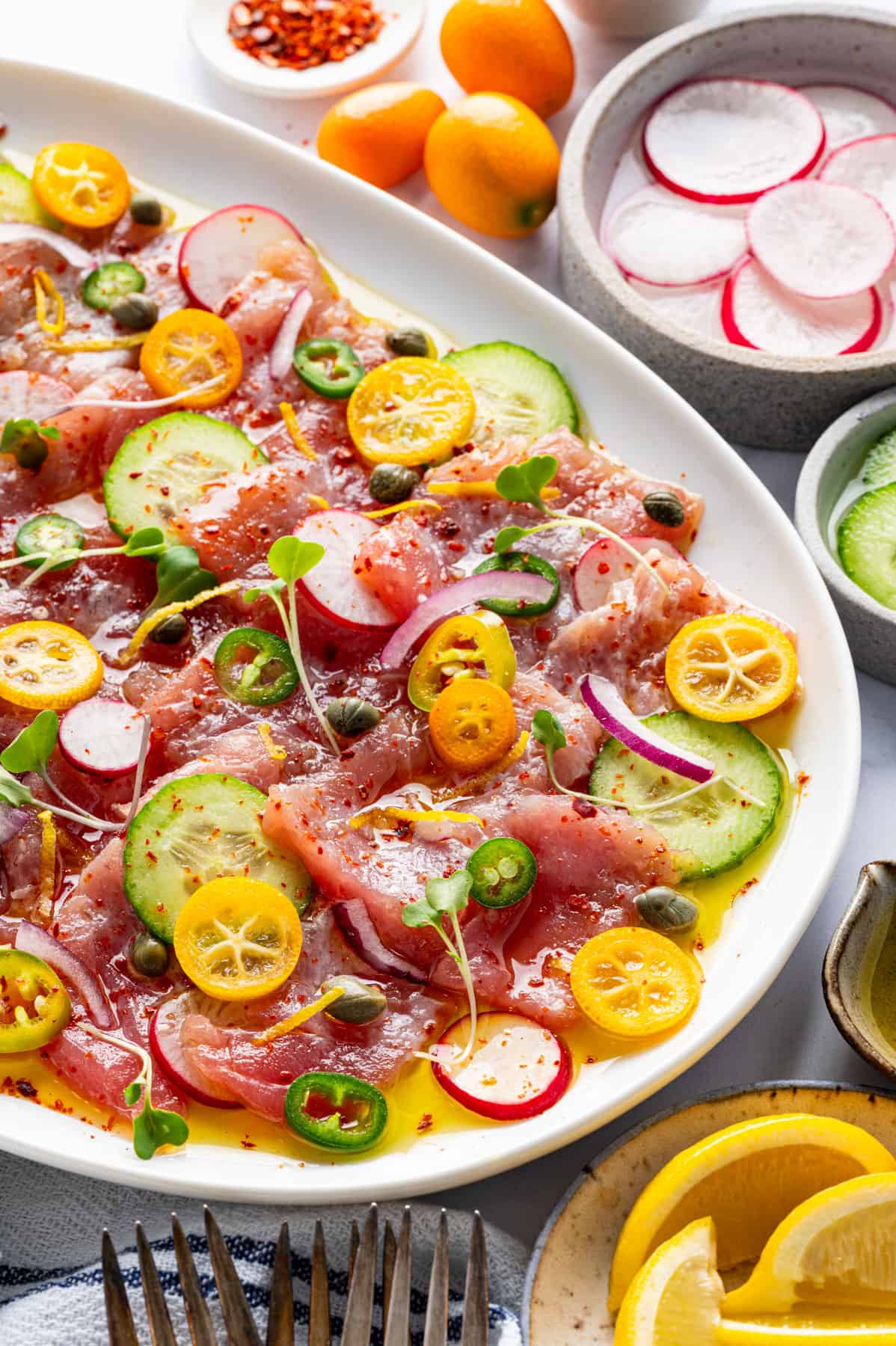 Tuna Crudo A platter of tuna crudo topped with sliced cucumber, radish, kumquats, jalapeños, capers, and red onions, drizzled with olive oil, with lemon wedges.