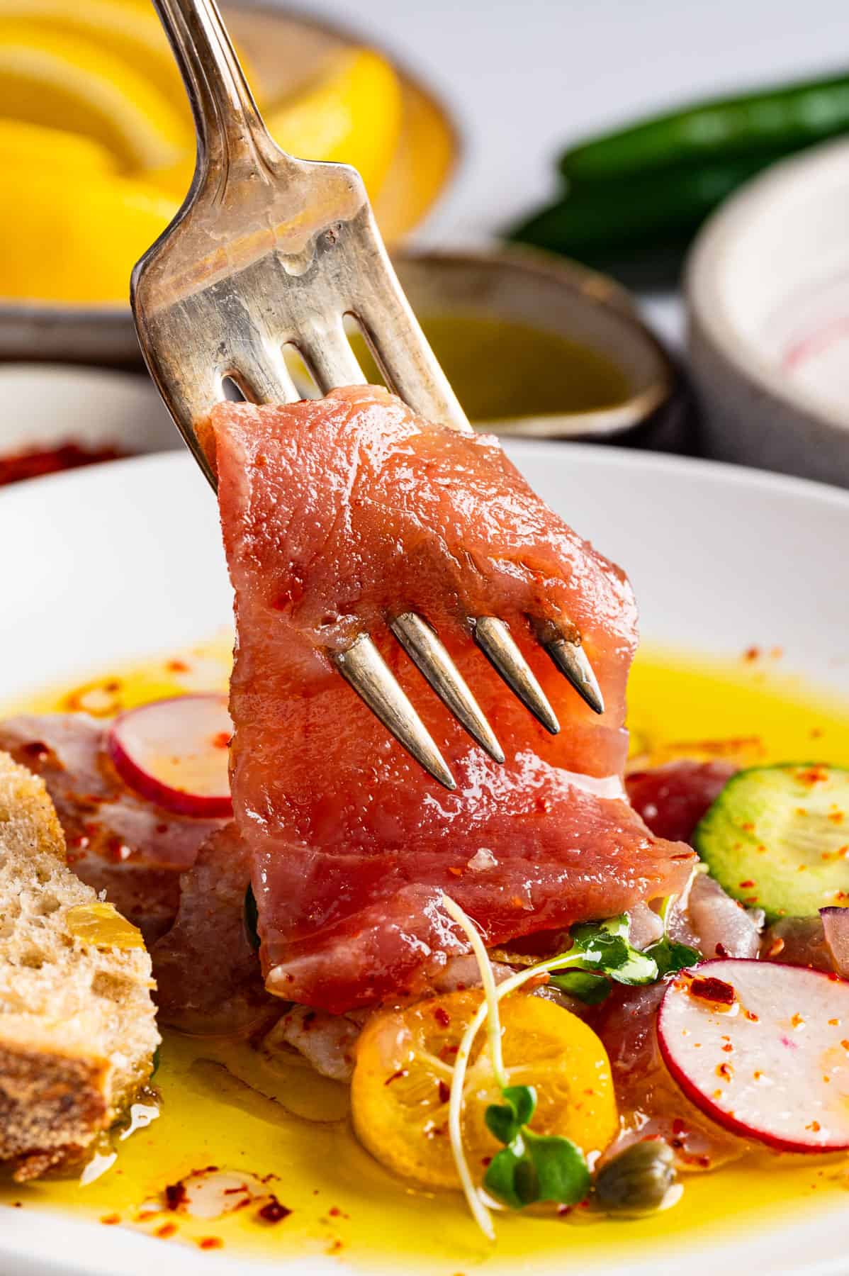 Tuna Crudo A fork holds a slice of raw tuna above a plate with yellow sauce, radish slices, cherry tomatoes, cucumbers, capers, and a piece of bread.