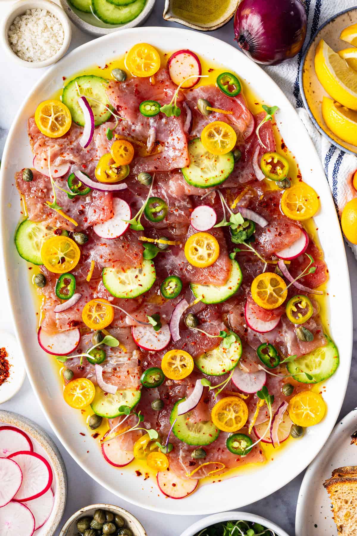 Tuna Crudo Oval platter of thinly sliced raw fish topped with cucumber, kumquat, red onion, jalapeño, capers, microgreens, and drizzled dressing.