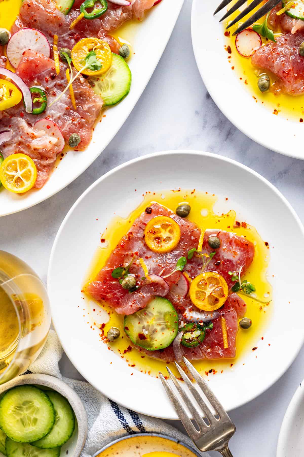 Tuna Crudo Plates of tuna crudo topped with capers, thinly sliced cucumbers, kumquats, chili flakes, onions, and olive oil.