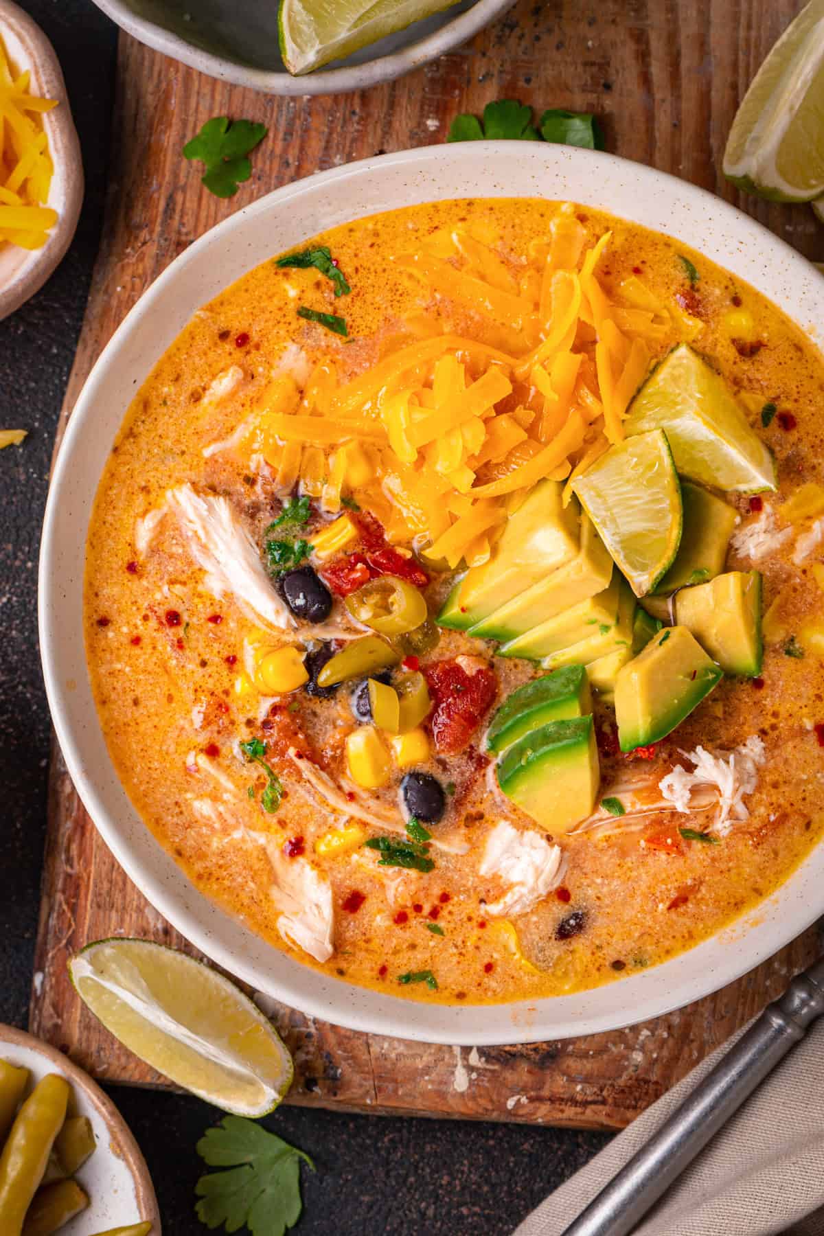 Chicken Enchilada Soup with corn, black beans, green chiles, topped with sliced avocado, Cheddar cheese, and sliced lime - in a bowl.