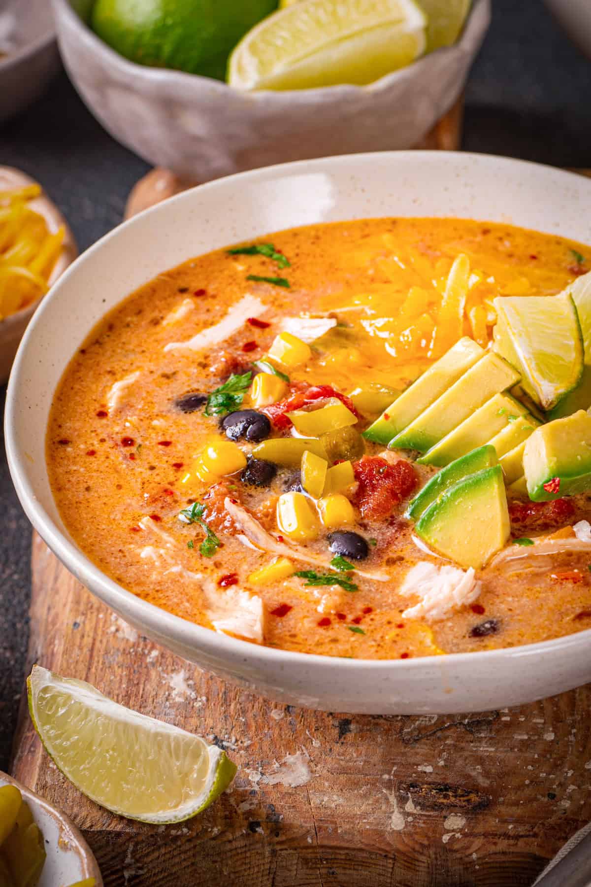 Chicken Enchilada Soup with corn, black beans, green chiles, topped with sliced avocado, Cheddar cheese, and sliced lime - in a bowl.
