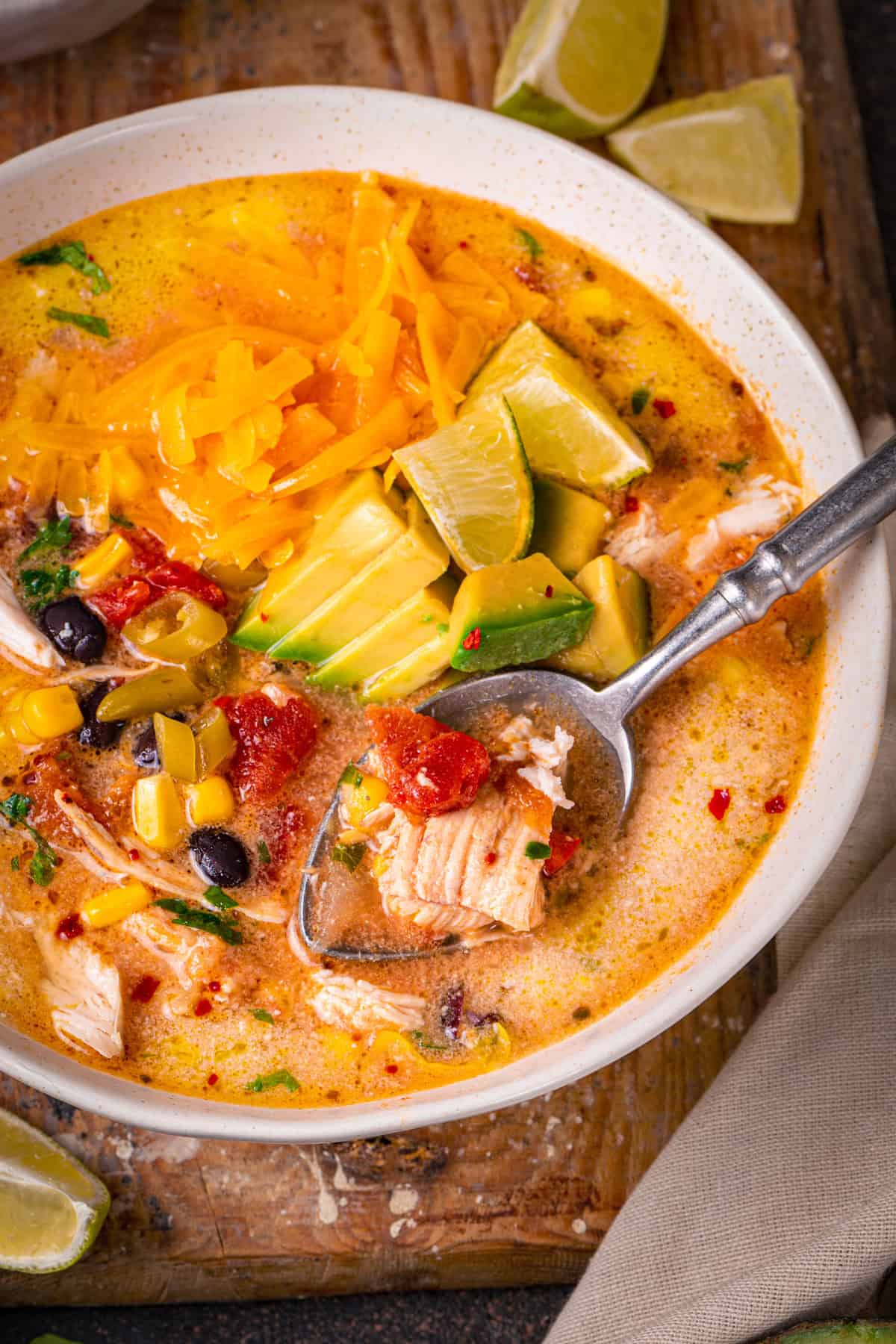 Chicken Enchilada Soup with corn, black beans, green chiles, topped with sliced avocado, Cheddar cheese, and sliced lime - in a bowl.