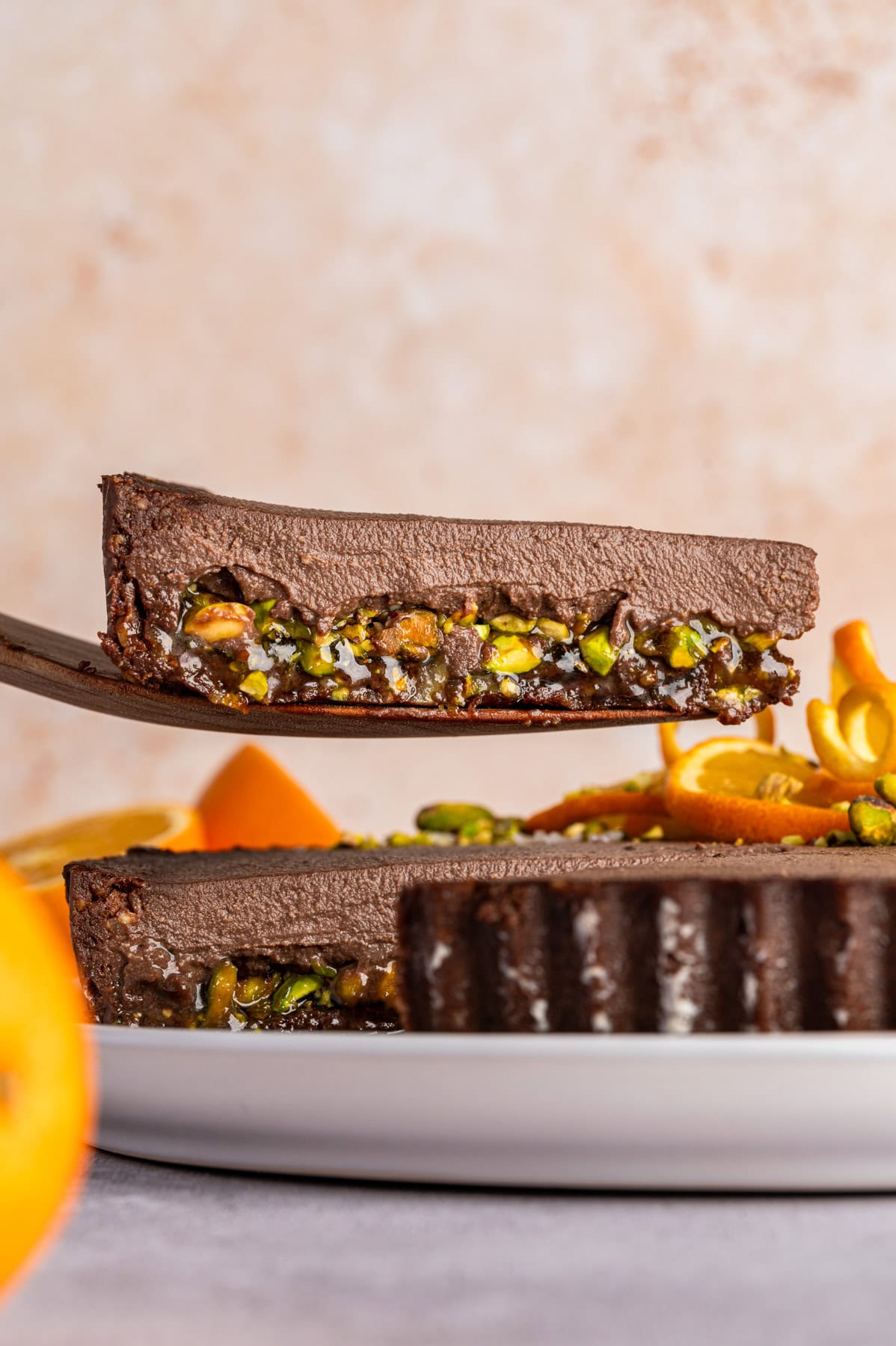 No-Bake Chocolate Tart with Orange Marmalade and Pistachios (Vegan + GF) A slice of Chocolate Orange Tart being lifted on a wooden spoon above the tart, showcasing the pistachio-orange marmalade and chocolate filling inside the tart.