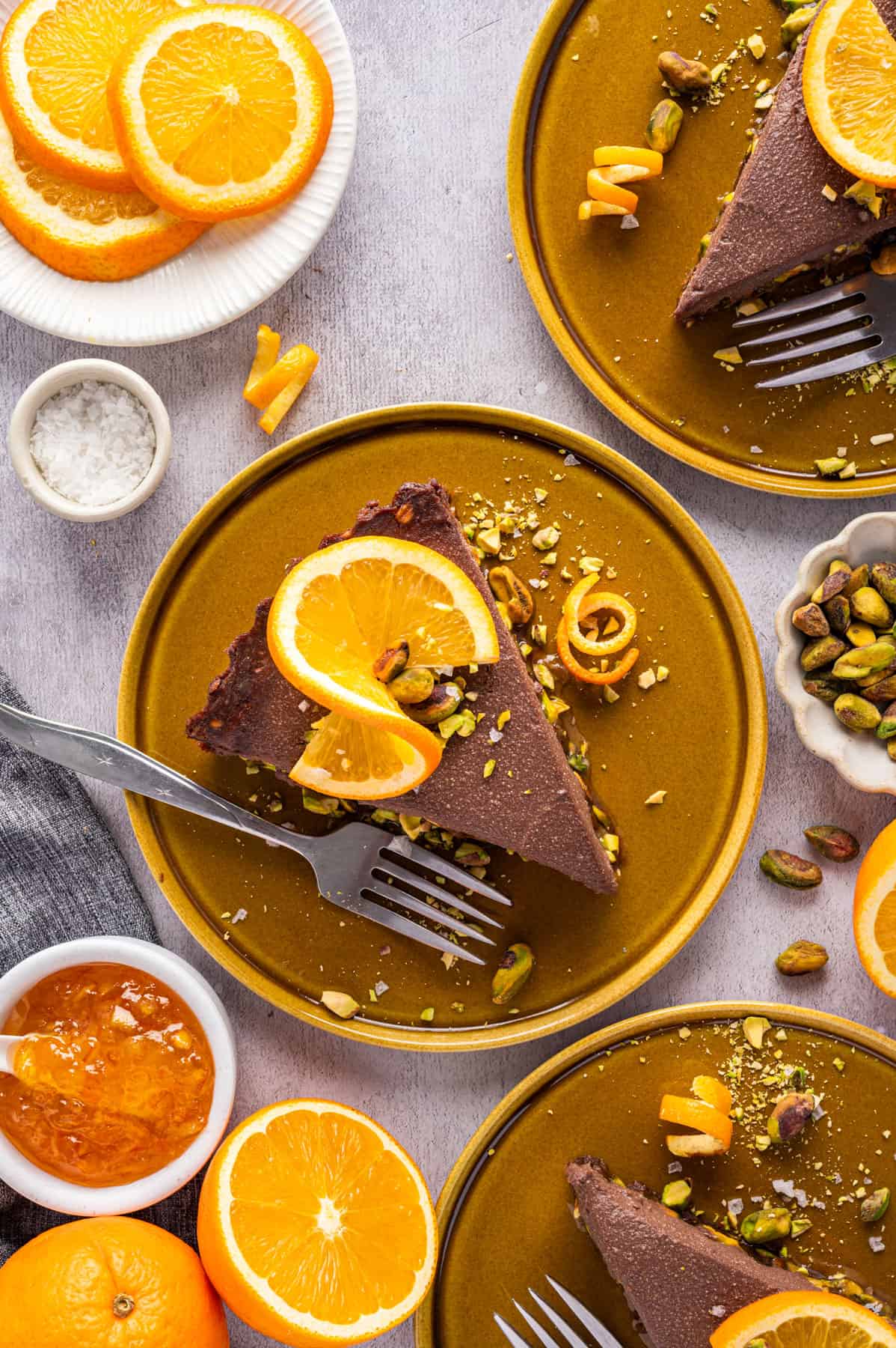 No-Bake Chocolate Tart with Orange Marmalade and Pistachios (Vegan + GF) A slice of Chocolate Orange Tart (topped with fresh orange slices, chopped and whole pistachios, and sea salt) on an orange plate surrounded by 2 more plates with tart slices.