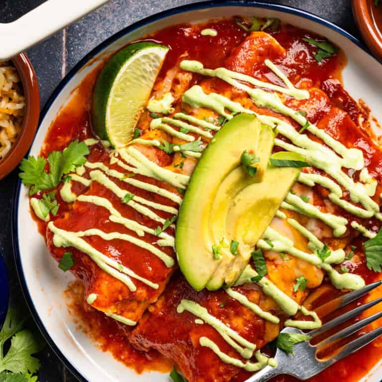 Chicken enchiladas with red enchilada sauce and drizzled with cilantro lime crema - on a white plate.