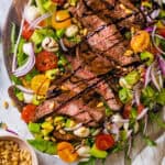 Grilled Steak Salad with baby greens, arugula, cherry tomatoes, red onions, mozzarella pearls, avocado, pine nuts, drizzled with balsamic glaze - on a large white plate.