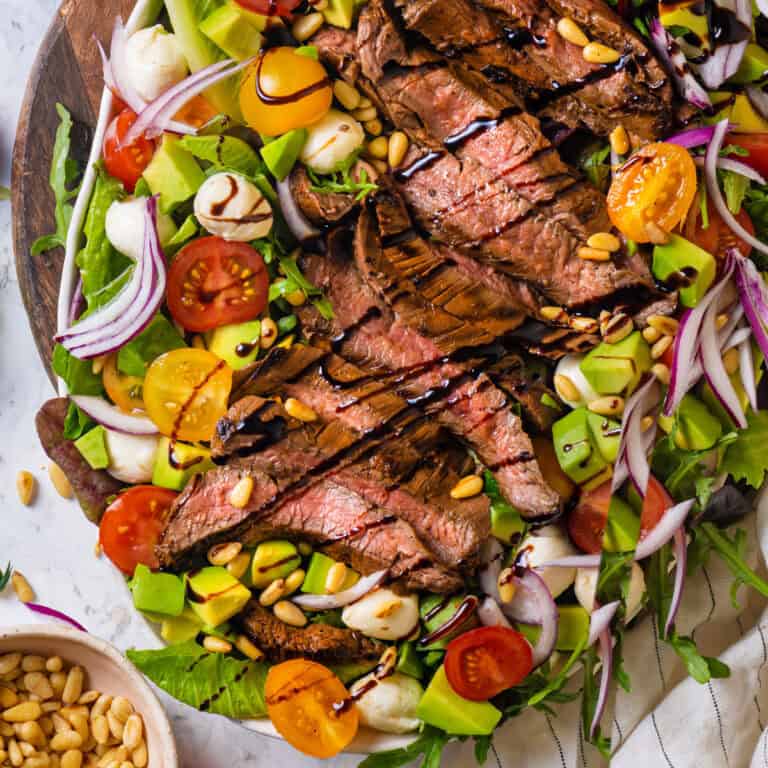Grilled Steak Salad with baby greens, arugula, cherry tomatoes, red onions, mozzarella pearls, avocado, pine nuts, drizzled with balsamic glaze - on a large white plate.