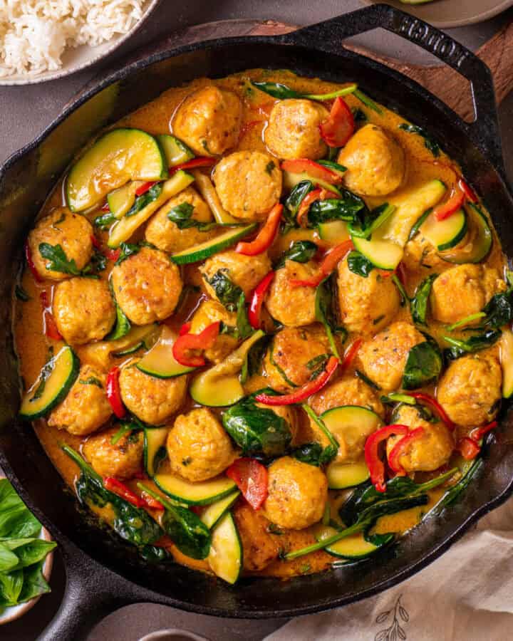 Thai chicken meatballs in a coconut milk red curry sauce with red bell pepper, zucchini, and spinach - in a cast iron skillet.