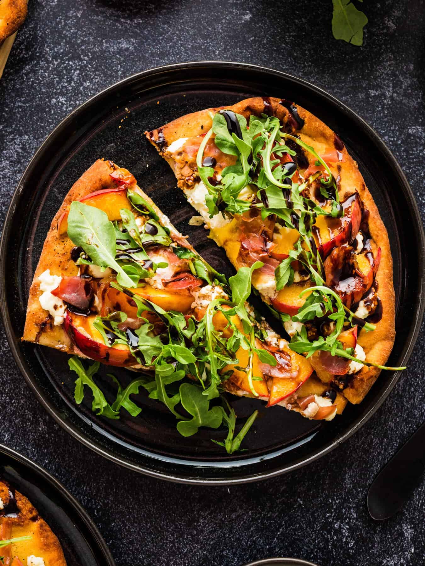 Peach Flatbread 2 slices of Peach Flatbread (with Prosciutto, Arugula, Goat Cheese, and Balsamic Glaze) - on a black plate.