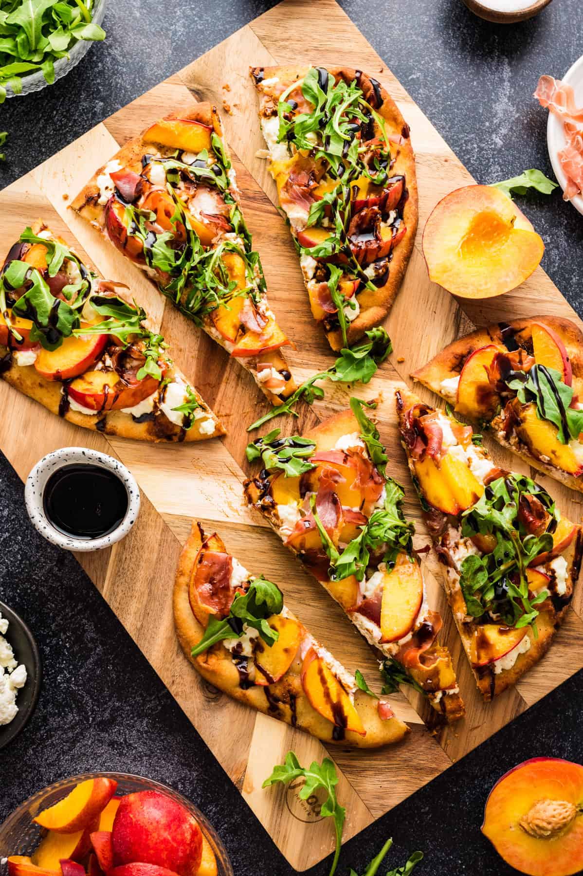Peach Flatbread Sliced Peach Flatbread (with Prosciutto, Arugula, Goat Cheese, and Balsamic Glaze) - on a wooden cutting board.