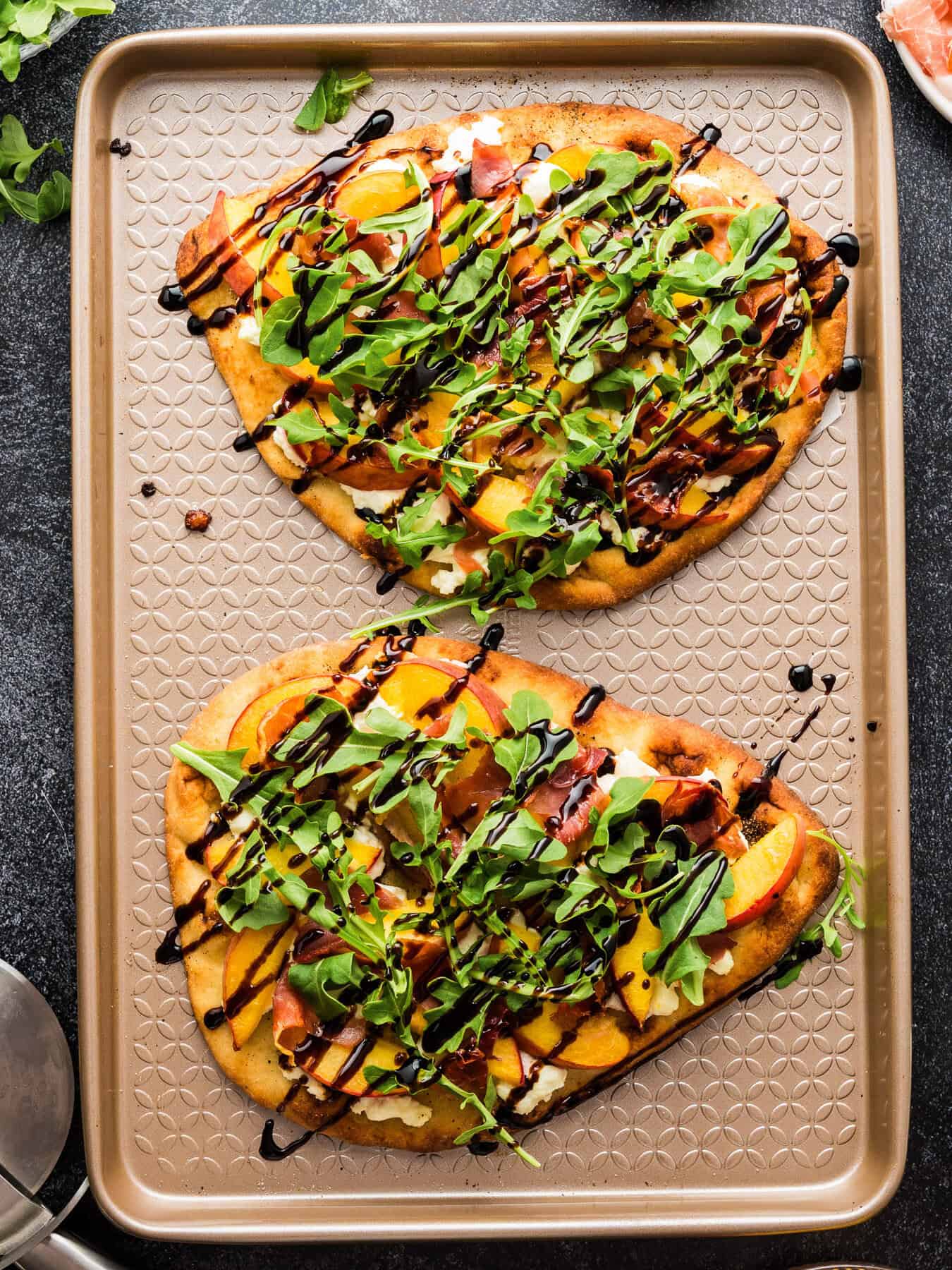 Peach Flatbread 2 unsliced whole Peach Flatbreads (with Prosciutto, Arugula, Goat Cheese, and Balsamic Glaze) - on a baking sheet.