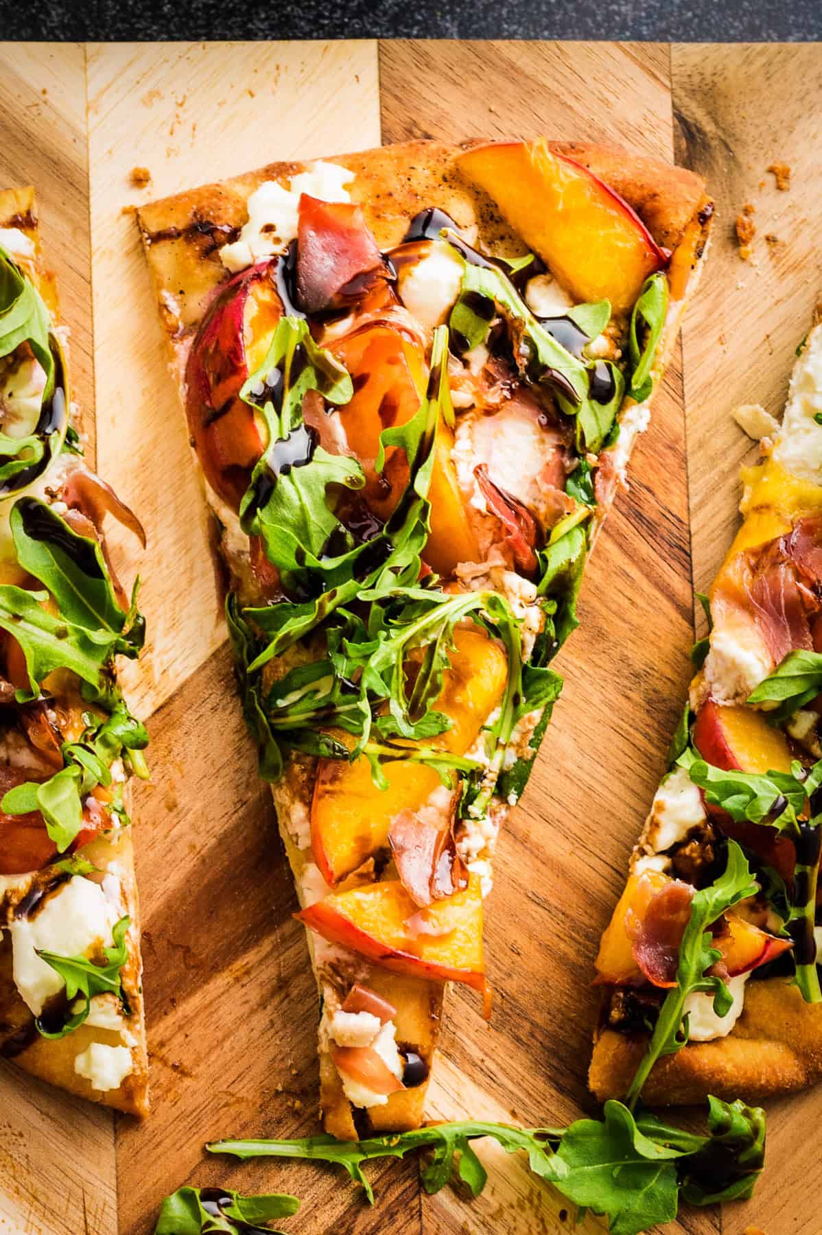 Peach Flatbread A slice of Peach Flatbread (with Prosciutto, Arugula, Goat Cheese, and Balsamic Glaze) - on a wooden cutting board.