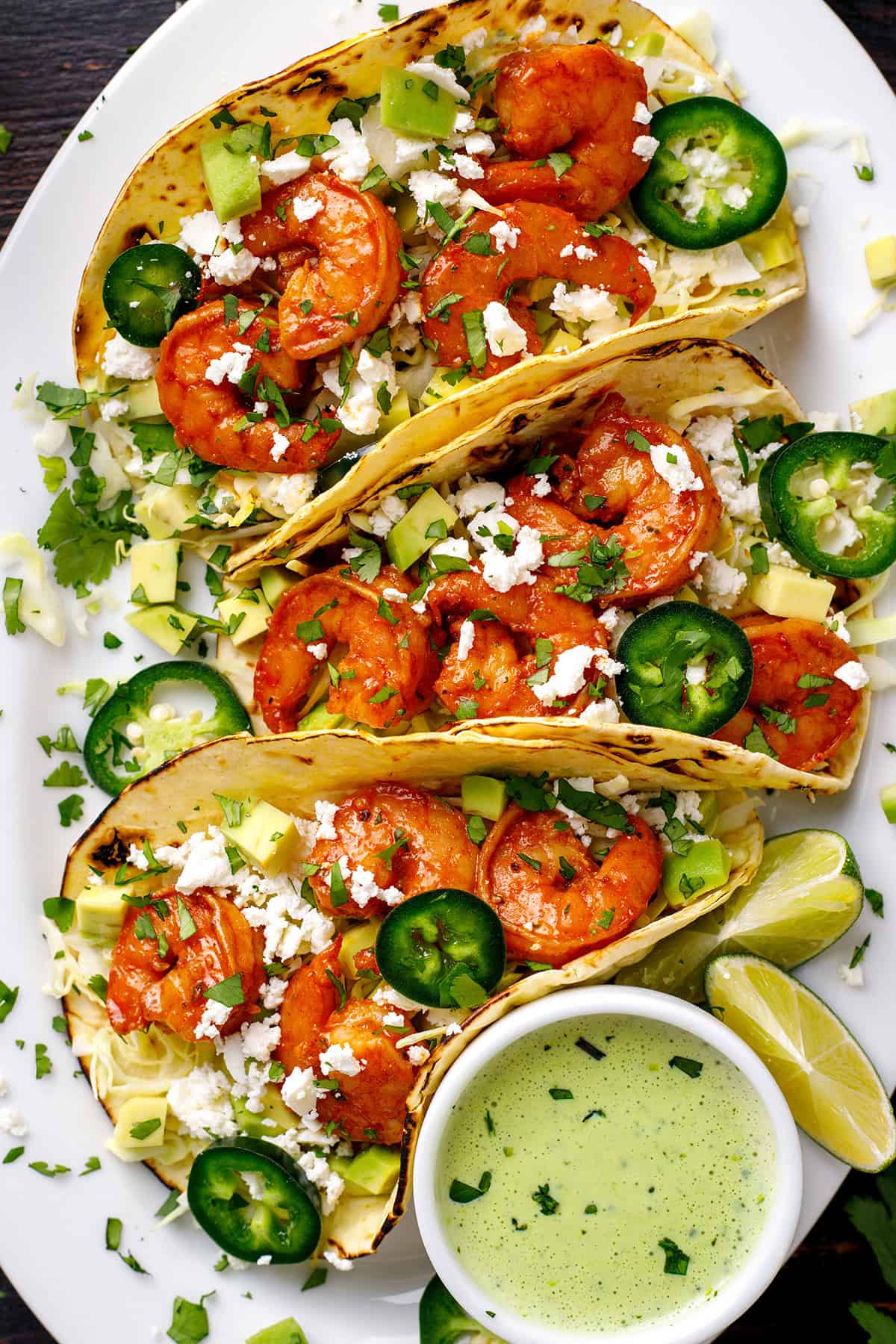 Shrimp Tacos Shrimp Tacos with shredded green cabbage, diced avocado, sliced jalapeños, crumbled feta cheese, and creamy cilantro sauce - on a white platter.