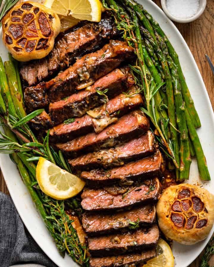 Seared Marinated Steak (thinly sliced) with asparagus and roasted garlic bulbs - on a large white platter.