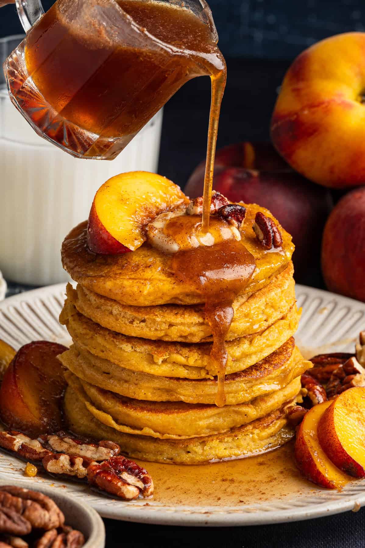 Peach Pancakes with Cinnamon Maple Syrup and Pecans A stack of pancakes with Cinnamon Maple Syrup being poured over the top, surrounded by peach slices, - on a white plate.