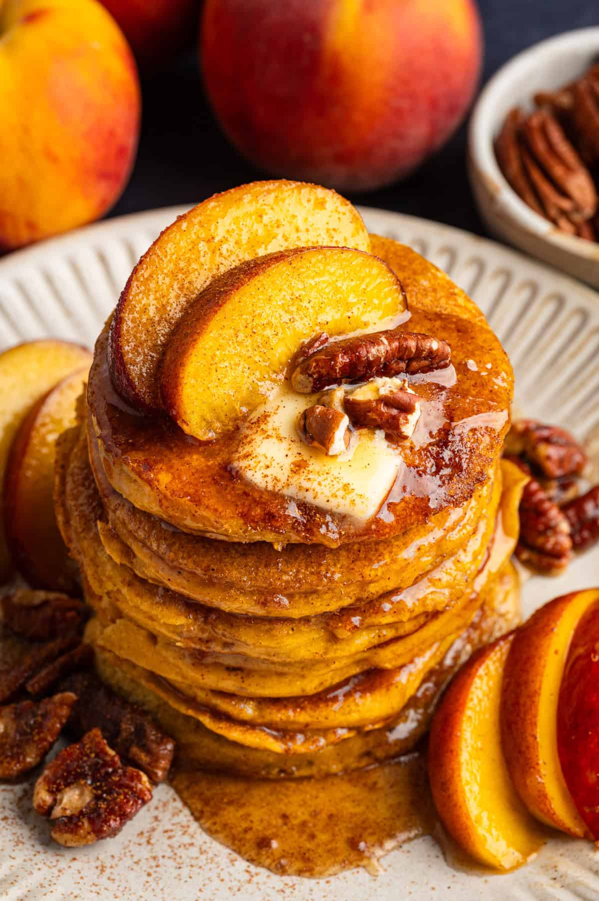 Peach Pancakes with Cinnamon Maple Syrup and Pecans A stack of Pancakes topped with Cinnamon Maple Syrup, Peach Slices, Butter, and Pecans - on a white plate.