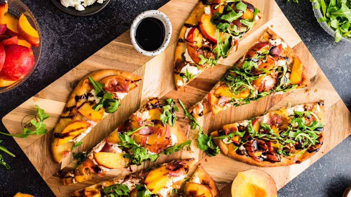 Peach, Goat Cheese & Prosciutto Flatbread (Your Summer Parties Need This!) Sliced Peach Flatbread (with Prosciutto, Arugula, Goat Cheese, and Balsamic Glaze) - on a wooden cutting board.