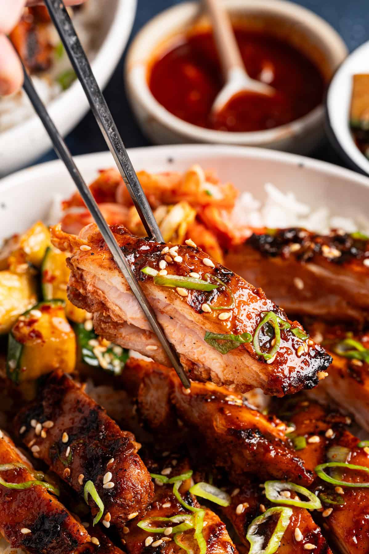 Korean BBQ Chicken with rice, kimchi, and sliced cucumber in a bowl (close up of a chicken slice being lifted by chopsticks).
