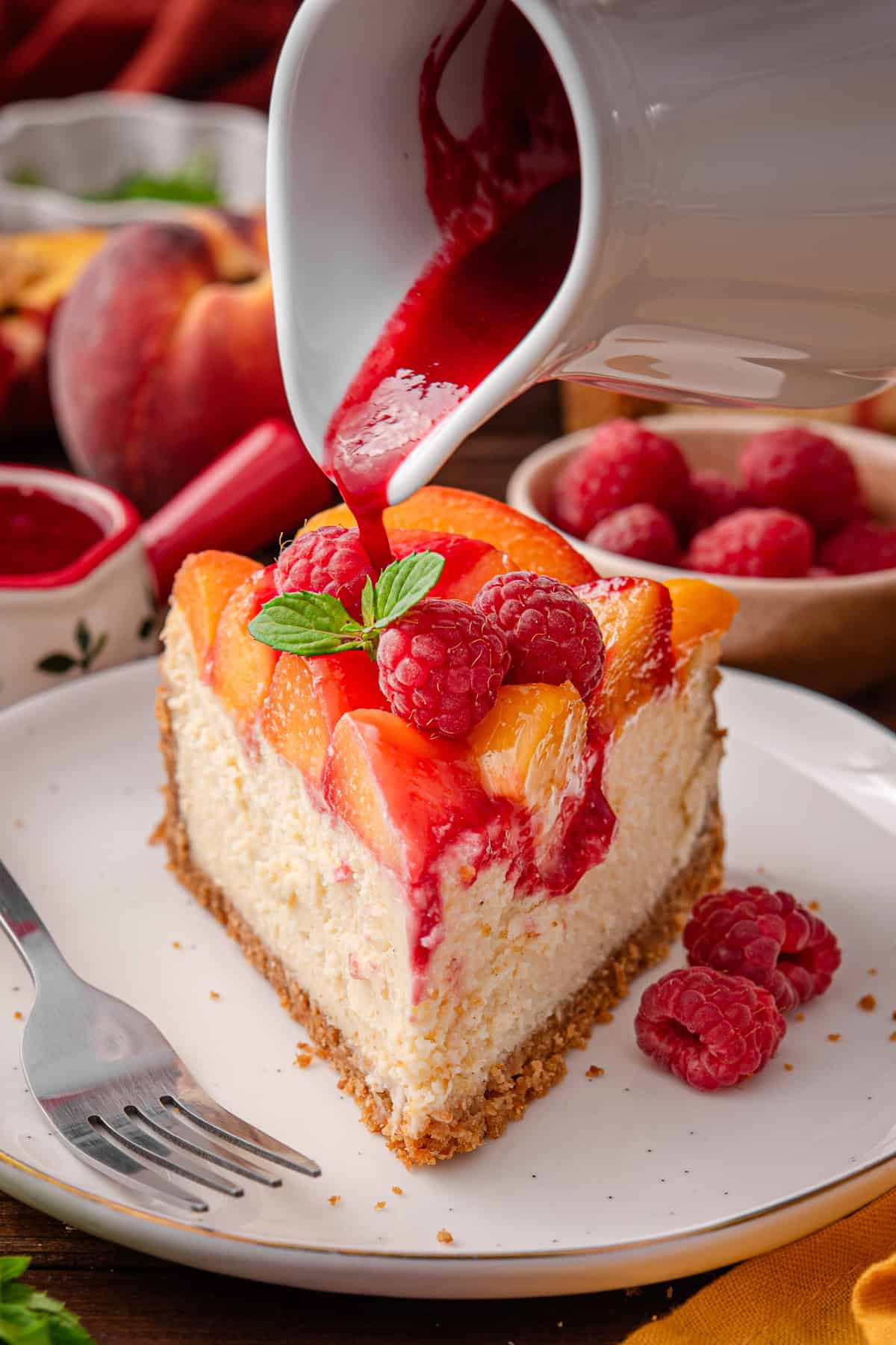 Peach Raspberry Cheesecake A slice of Peach Raspberry Cheesecake on a plate, with the raspberry sauce being poured on top of the slice.