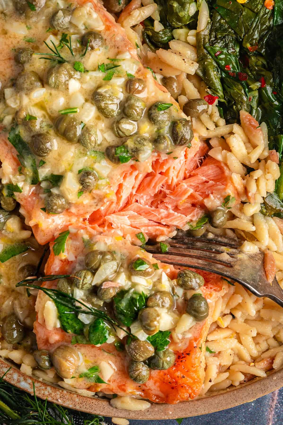 Salmon Piccata with Orzo and Spinach Close-up photo of Salmon Piccata - one salmon fillet topped with capers and plated with cooked orzo and spinach on a plate.