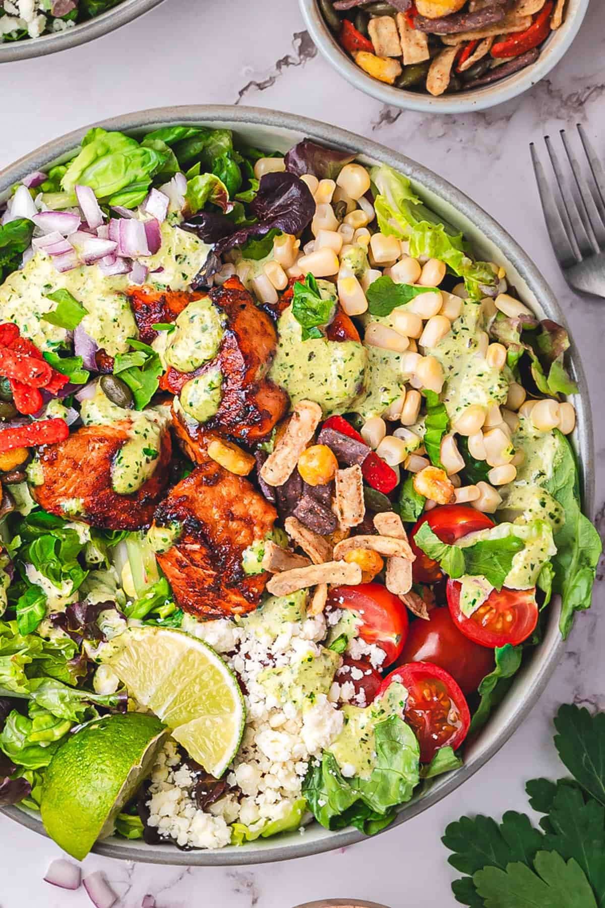 Mexican Salmon Salad Bowl with Roasted Poblano Ranch Salmon taco salad with mixed lettuce, corn, cherry tomatoes, red onion, Cotija cheese, lime wedges, fresh cilantro, and creamy poblano ranch dressing in a bowl.