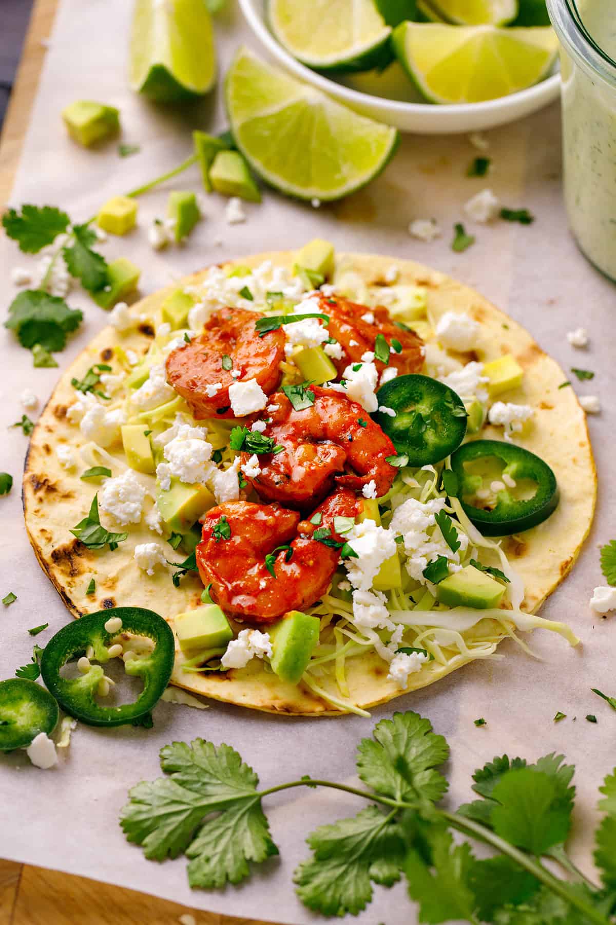 Shrimp Tacos A single Shrimp Taco with shredded green cabbage, diced avocado, sliced jalapeños, crumbled feta cheese - on a white surface.
