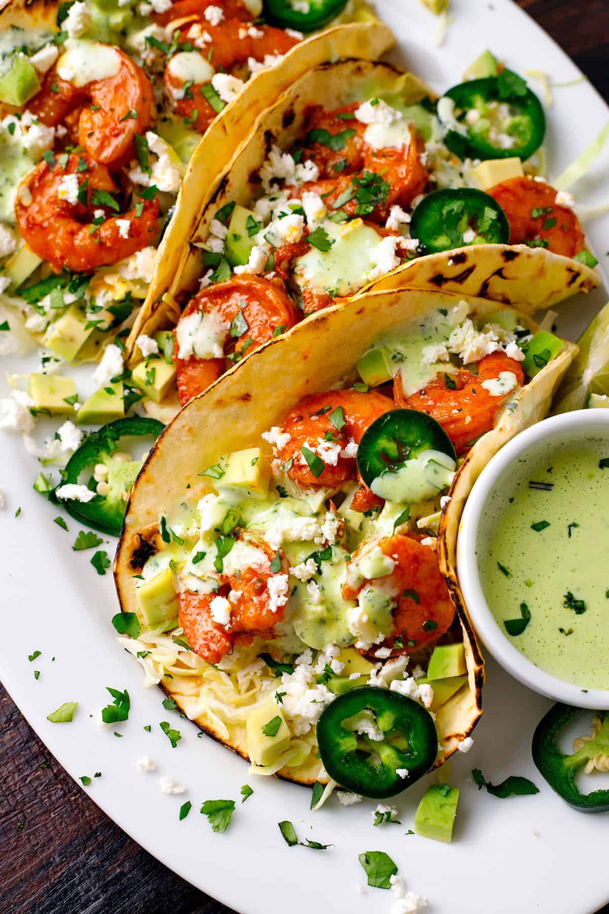 Shrimp Tacos Shrimp Tacos with shredded green cabbage, diced avocado, sliced jalapeños, crumbled feta cheese, and creamy cilantro sauce - on a white platter.