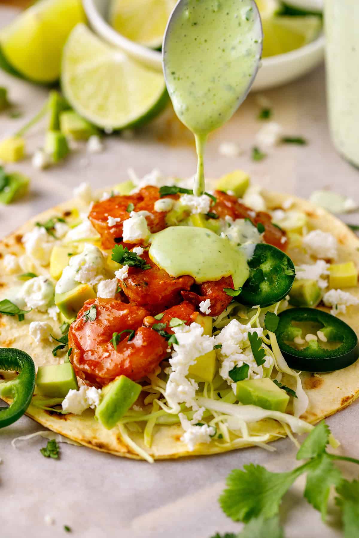 Shrimp Tacos A single Shrimp Taco topped with shredded green cabbage, diced avocado, sliced jalapeños, crumbled feta cheese, and with the light green creamy cilantro sauce being poured on top.