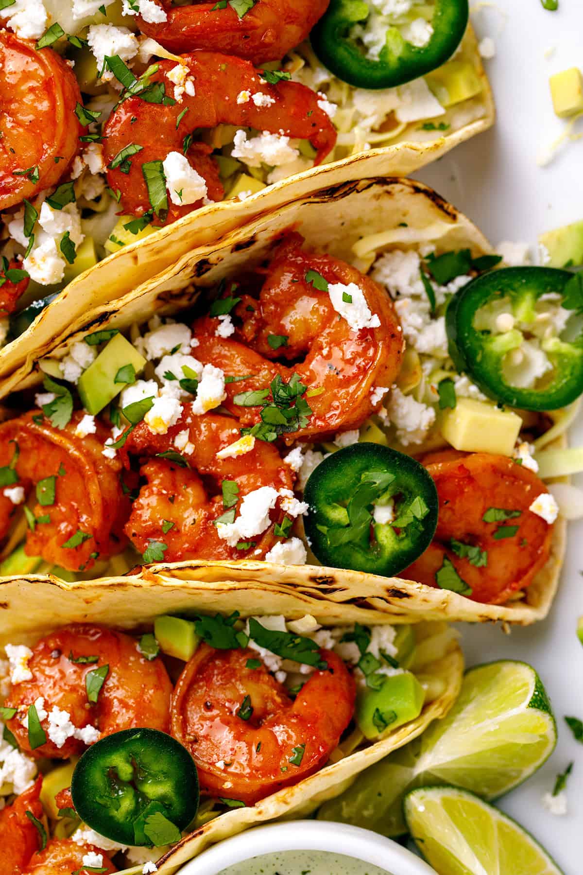 Shrimp Tacos Shrimp Tacos with shredded green cabbage, diced avocado, sliced jalapeños, crumbled feta cheese - on a white platter.