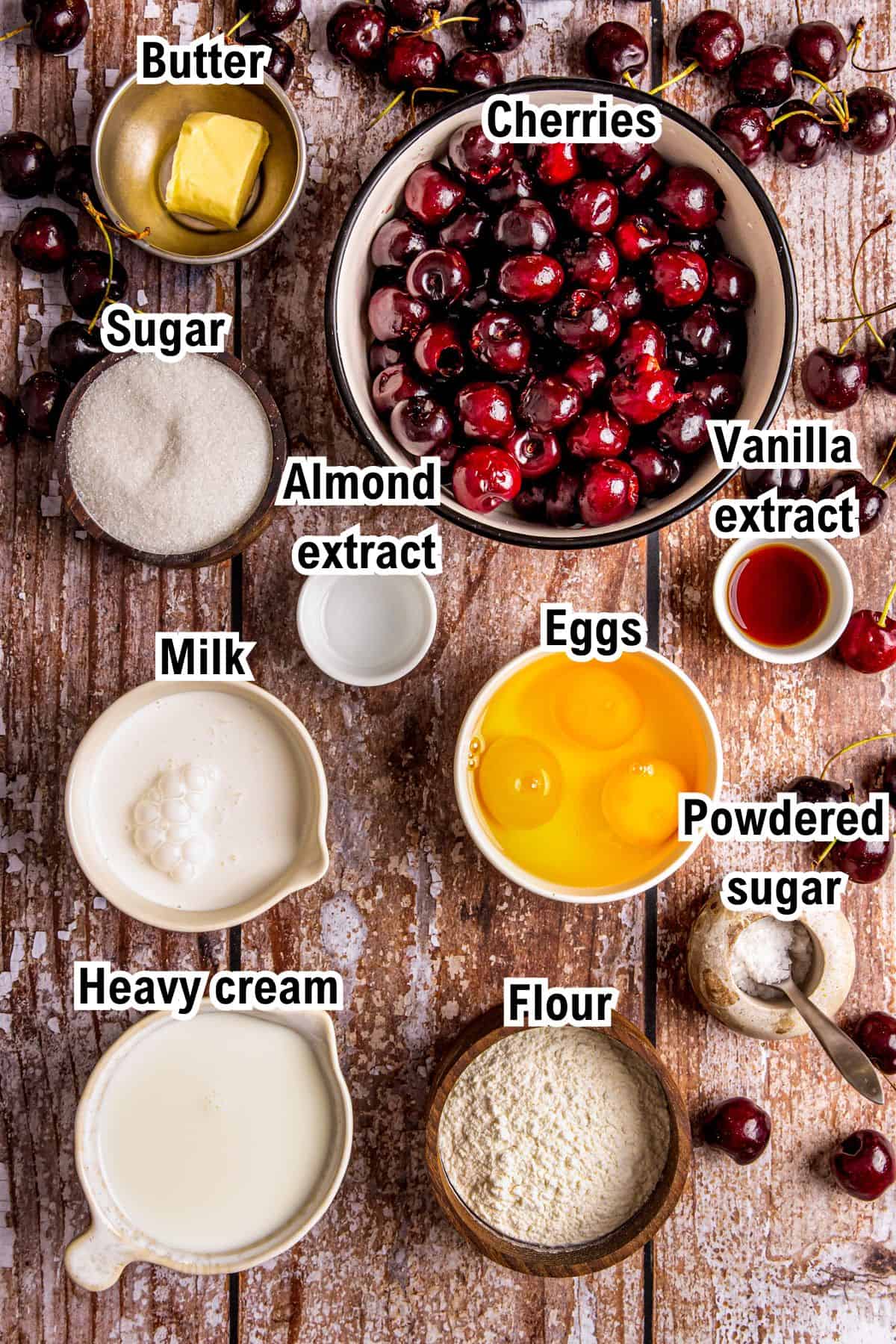 A wooden table displays the ingredients for Cherry Clafoutis, including labeled bowls containing cherries, butter, sugar, almond extract, milk, heavy cream, flour, eggs, vanilla extract, and powdered sugar, arranged for baking.
