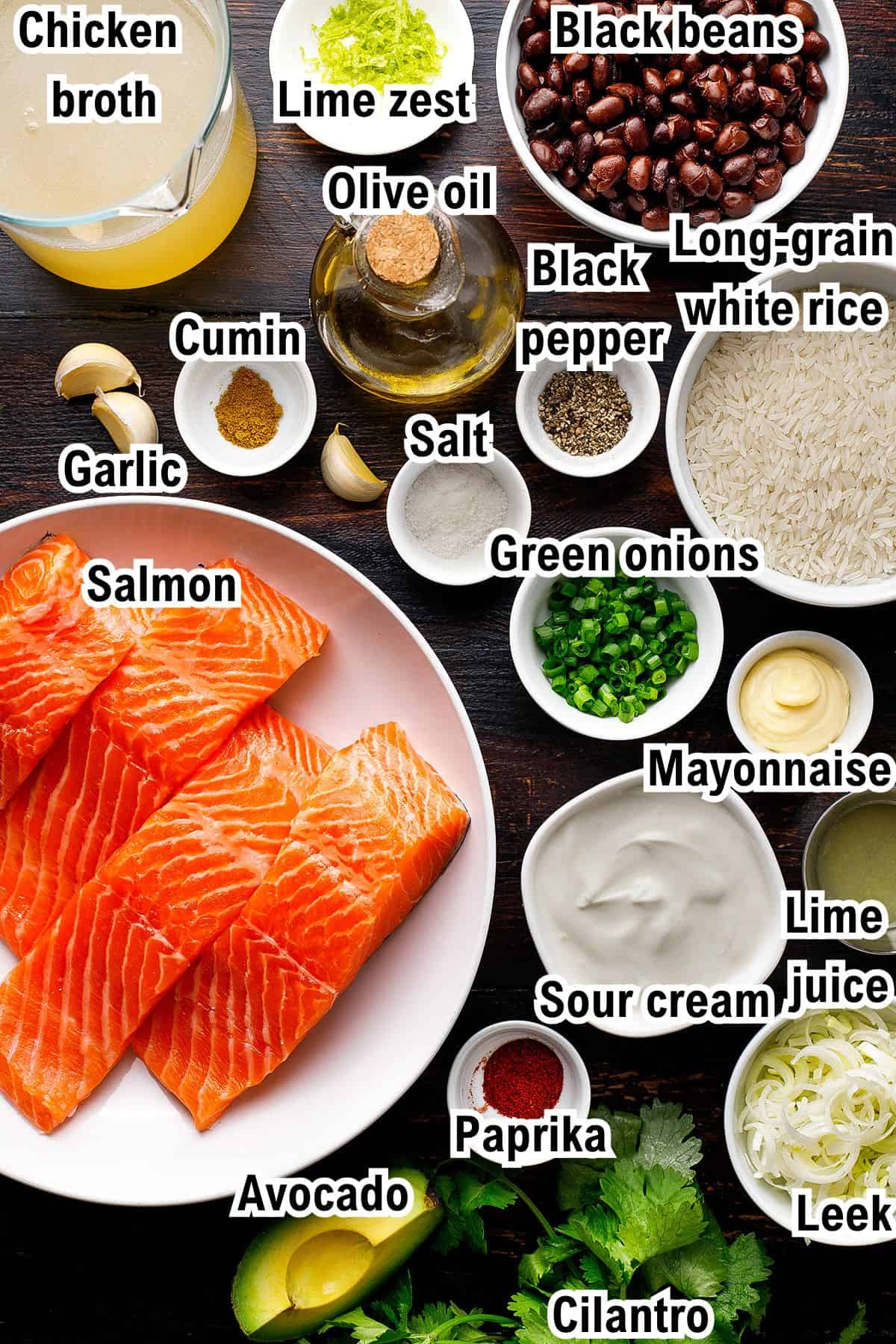Top view of various labeled ingredients for Salmon and Rice on a dark surface, including raw salmon fillets, avocado, mayonnaise, sour cream, lime, black beans, white rice, chicken broth, spices, olive oil, garlic, leeks, green onions, and cilantro.