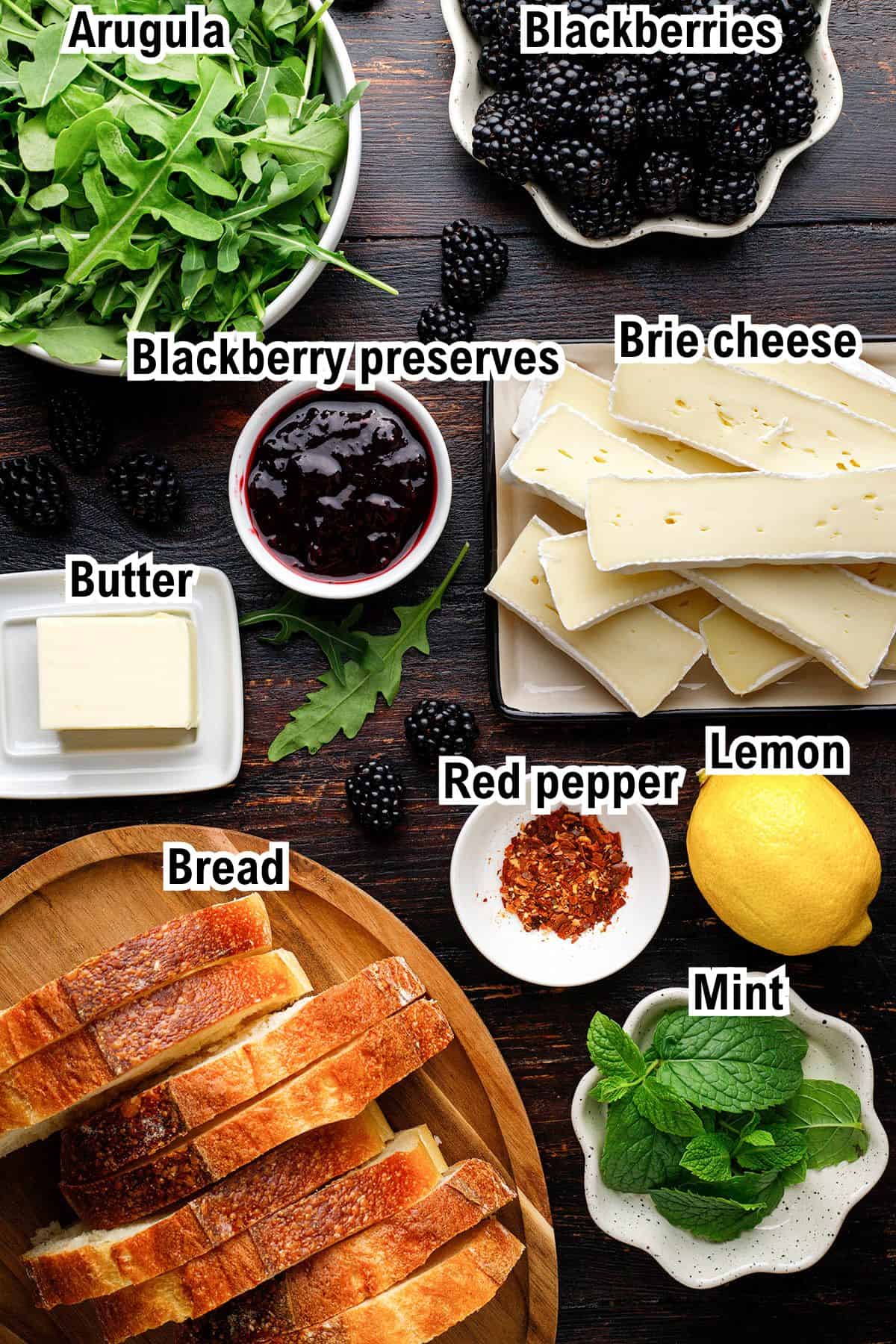 A flat lay of ingredients for Blackberry Brie Grilled Cheese Sandwich: arugula, blackberries, blackberry preserves, Brie cheese slices, butter, sliced bread, red pepper flakes, a lemon, and mint leaves, all labeled with text.