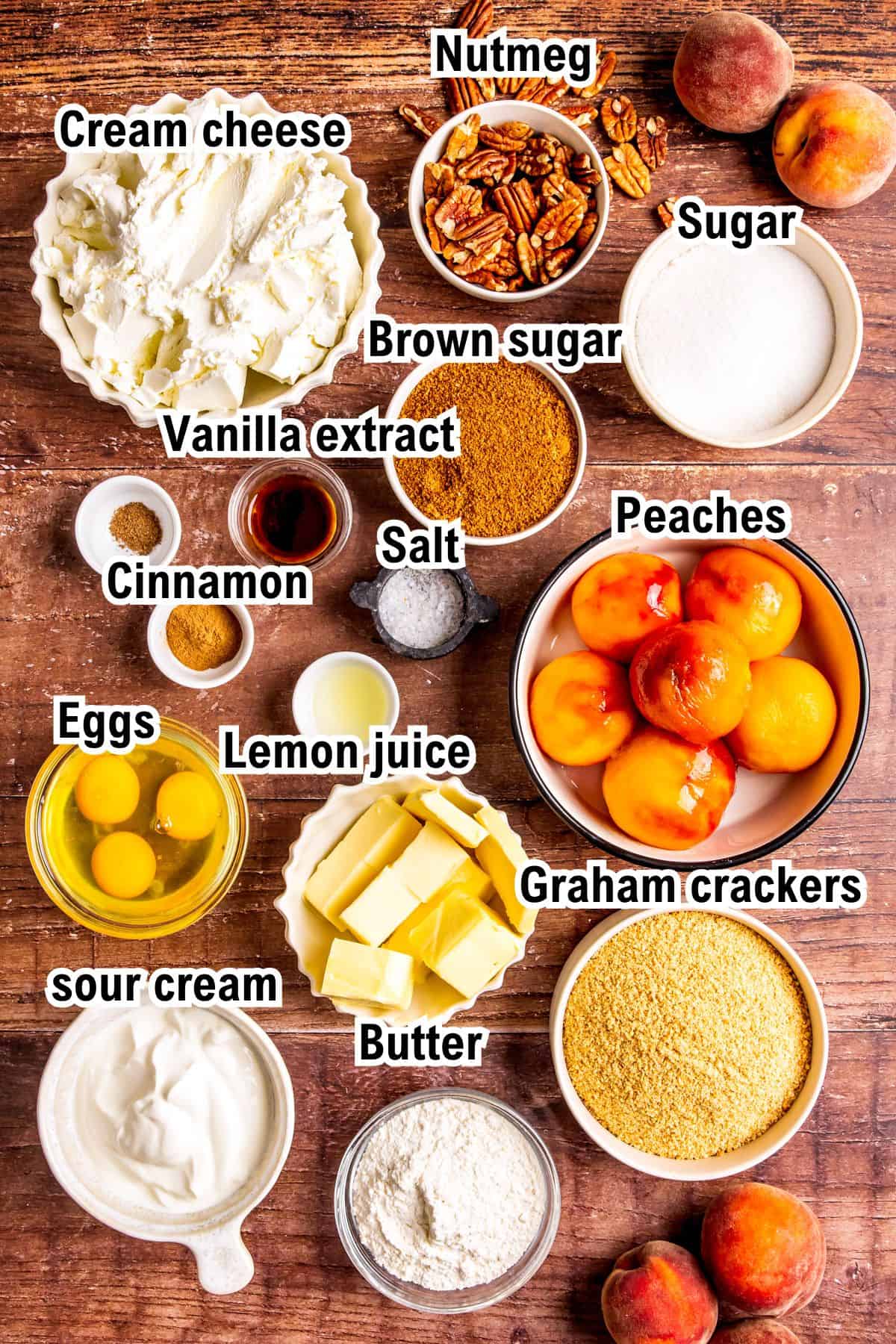 A variety of baking ingredients for Peach Cobbler Cheesecake on a wooden surface, labeled: cream cheese, nutmeg, pecans, brown sugar, sugar, vanilla extract, cinnamon, salt, peaches, eggs, lemon juice, graham crackers, butter, and sour cream.