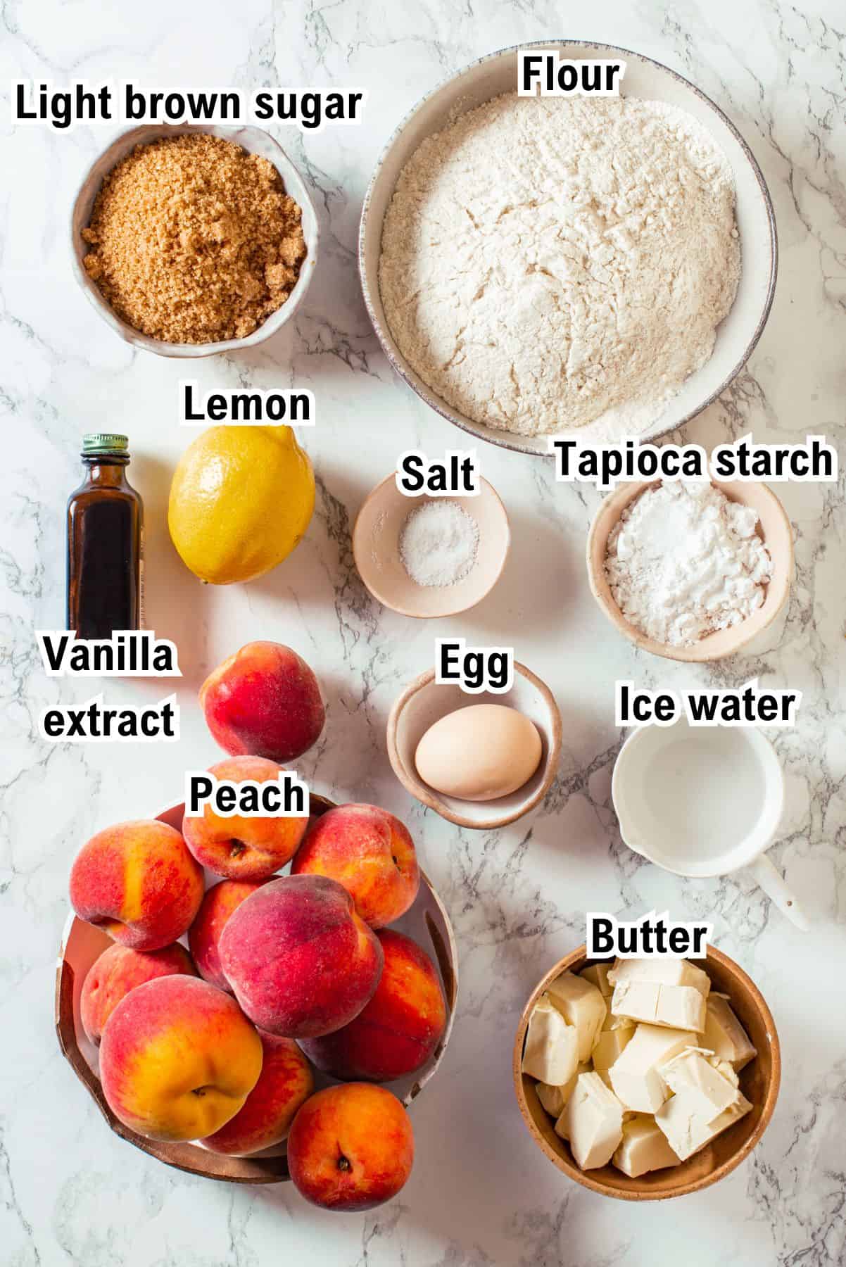 Overhead view of baking ingredients for Peach Pie on a marble surface, labeled: light brown sugar, flour, lemon, salt, tapioca starch, vanilla extract, egg, ice water, butter, and several fresh peaches.