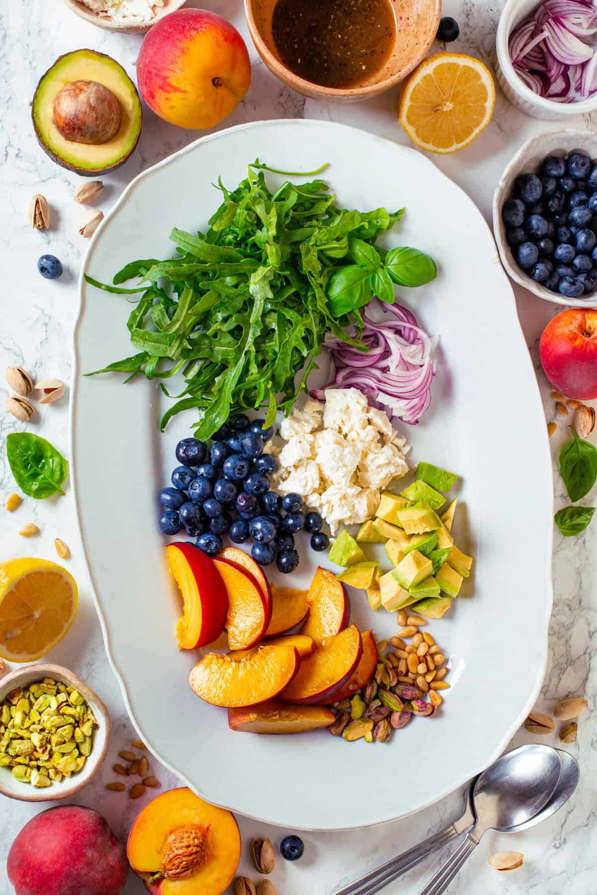 The Ingredients for Grilled Peach Salad The Salad Ingredients: sliced Peaches, Arugula, Blueberries, Feta, Avocado, Pine Nuts, and Pistachios - on a large white platter.