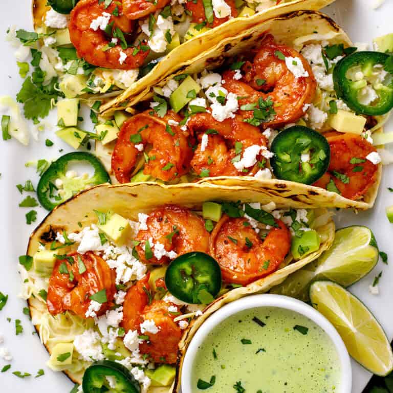 Shrimp Tacos with shredded green cabbage, diced avocado, sliced jalape&ntilde;os, crumbled feta cheese, and creamy cilantro sauce - on a white platter.