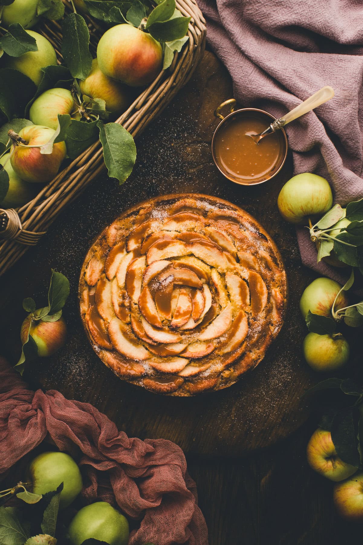 German Apple Cake (drizzled with caramel sauce( on a dark background surrounded by apples and a basket with apples.