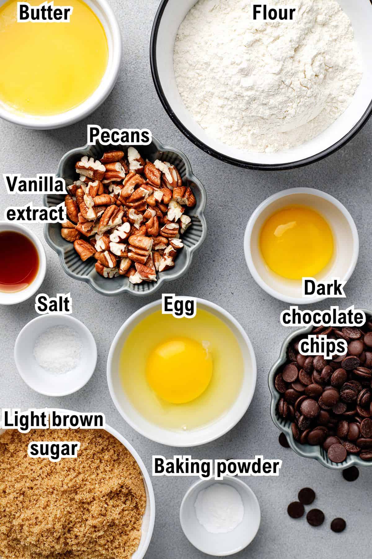 The Ingredients for Blondies Bowls with labeled ingredients for blondies: butter, flour, pecans, vanilla extract, dark chocolate chips, salt, light brown sugar, egg, and baking powder, arranged on a light surface.