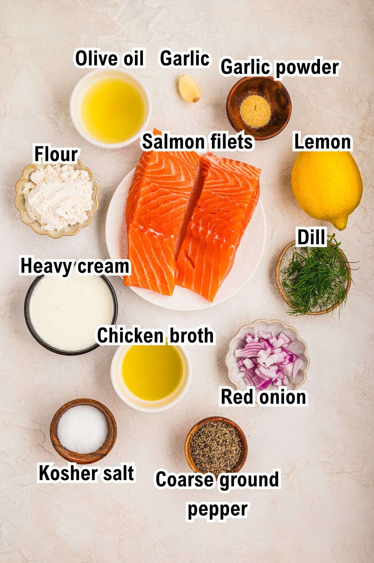 Two raw salmon fillets on a plate surrounded by labeled ingredients: olive oil, garlic, garlic powder, lemon, dill, red onion, chicken broth, heavy cream, flour, kosher salt, and coarse ground pepper.