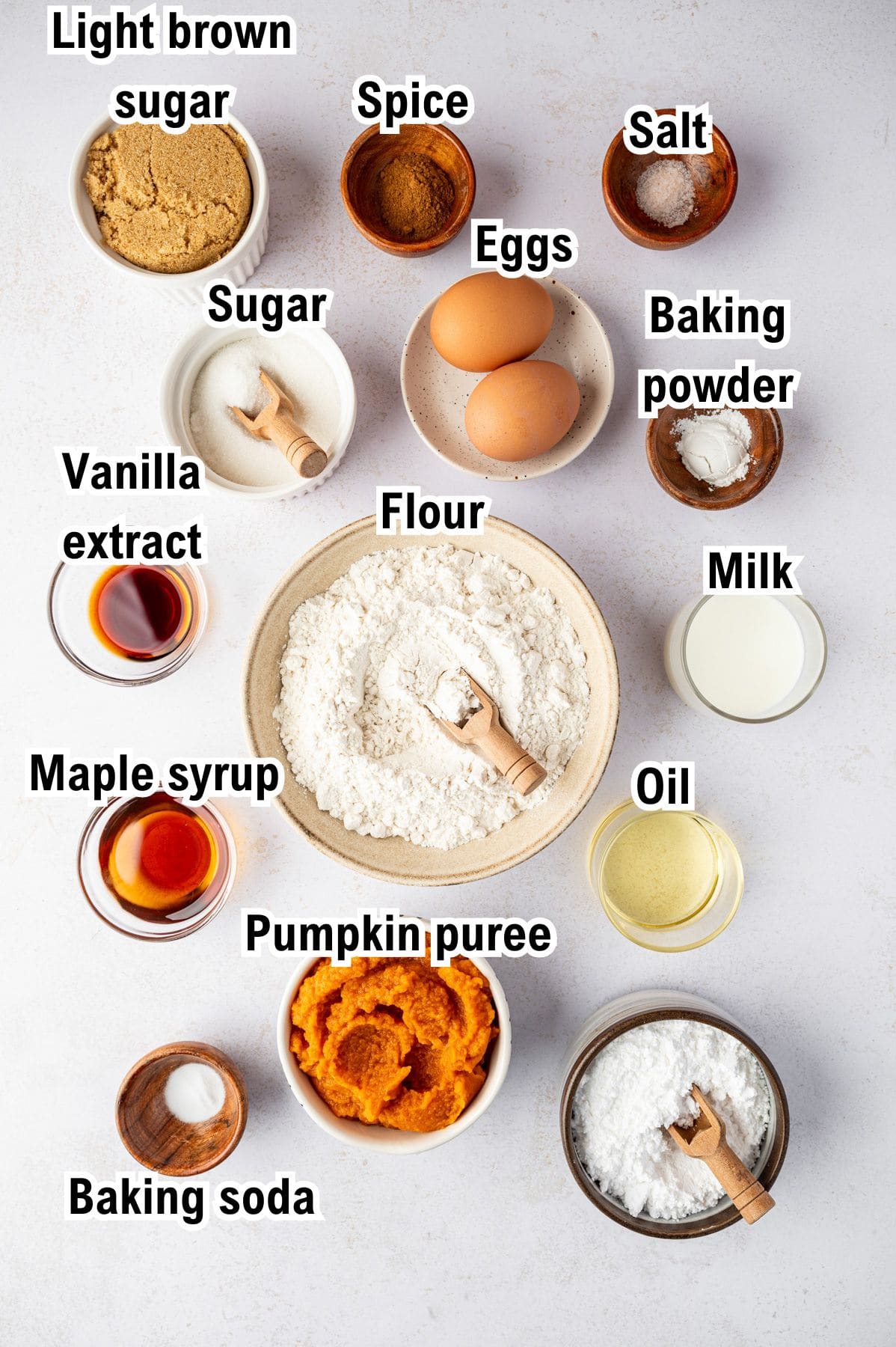 The Ingredients for Pumpkin Spice Donuts This photo shows the ingredients for Pumpkin Spice Donuts: flour, eggs, milk, oil, pumpkin puree, maple syrup, vanilla extract, light brown sugar, sugar, spice, salt, baking powder, and baking soda in bowls on a white surface.