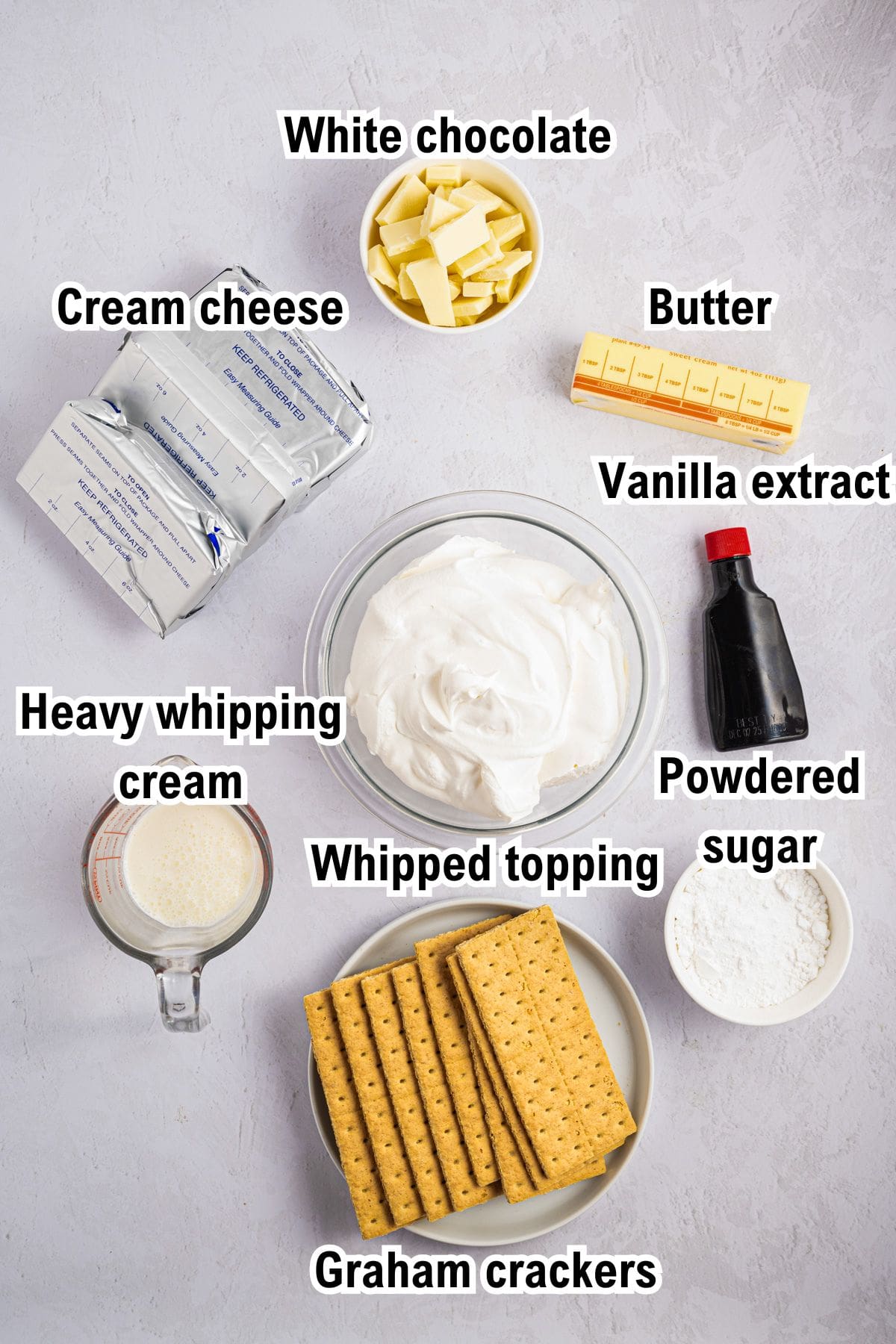 The Ingredients for White Chocolate Cheesecake This photo shows the ingredients for White Chocolate Cheesecake: White chocolate, cream cheese, butter, vanilla extract, heavy whipping cream, whipped topping, powdered sugar, and graham crackers arranged on a light background.