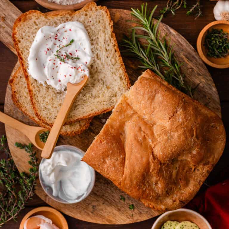 A loaf of Bread Machine Garlic Bread with one slice sliced off (and topped with a white spread) - on a dark wooden background.