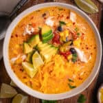 Chicken Enchilada Soup with corn, black beans, green chiles, topped with sliced avocado, Cheddar cheese, and sliced lime - in a bowl.