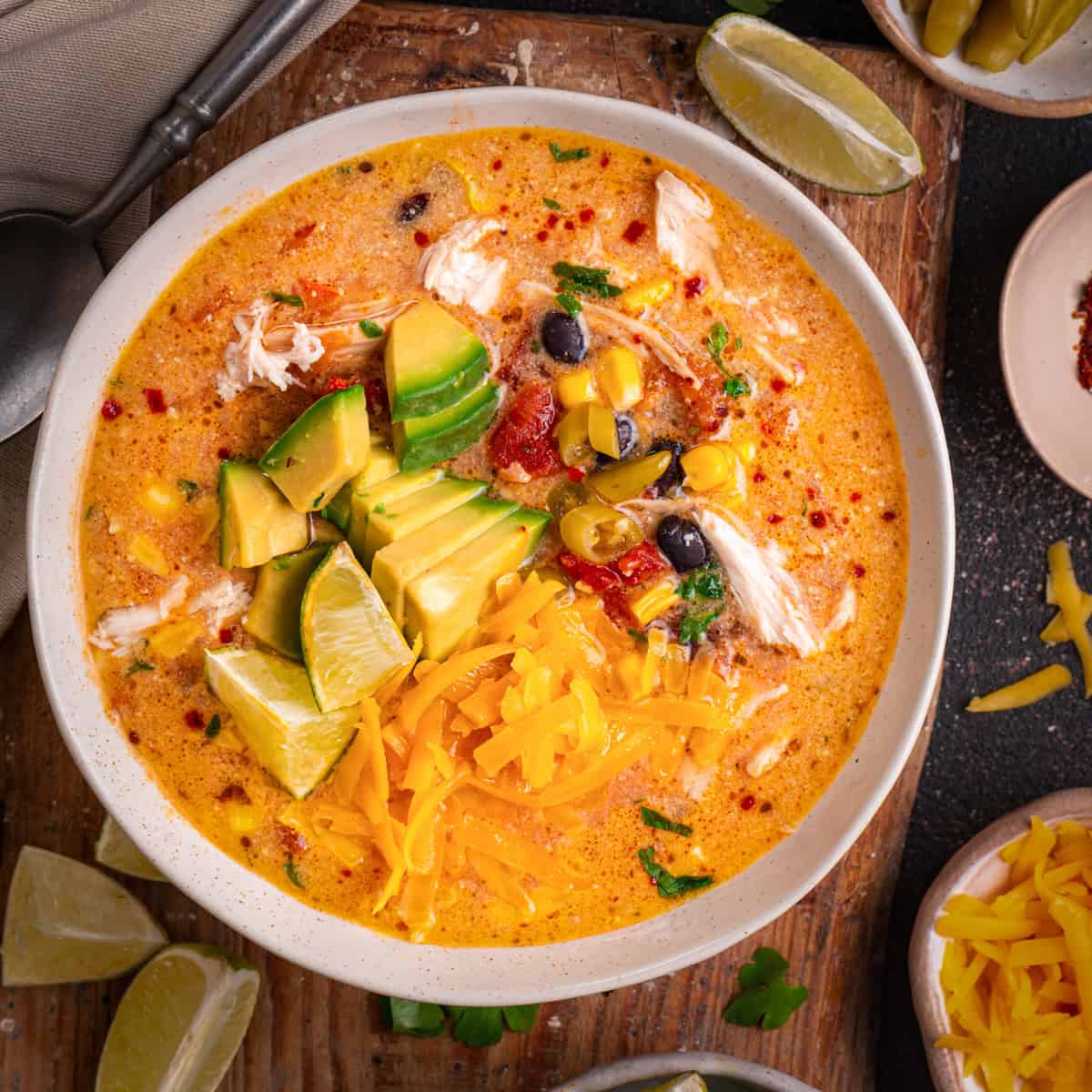Chicken Enchilada Soup with corn, black beans, green chiles, topped with sliced avocado, Cheddar cheese, and sliced lime - in a bowl.