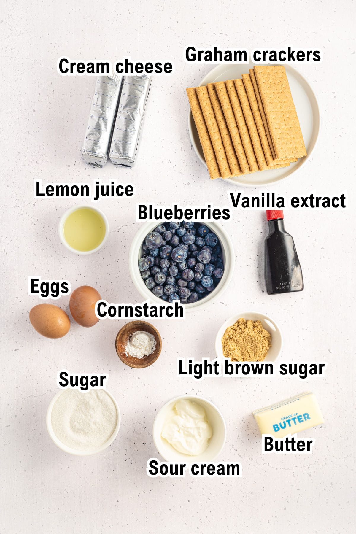 Top-down view of labeled blueberry cheesecake bars ingredients, including cream cheese, graham crackers, lemon juice, blueberries, vanilla extract, eggs, cornstarch, light brown sugar, sugar, sour cream, and butter.