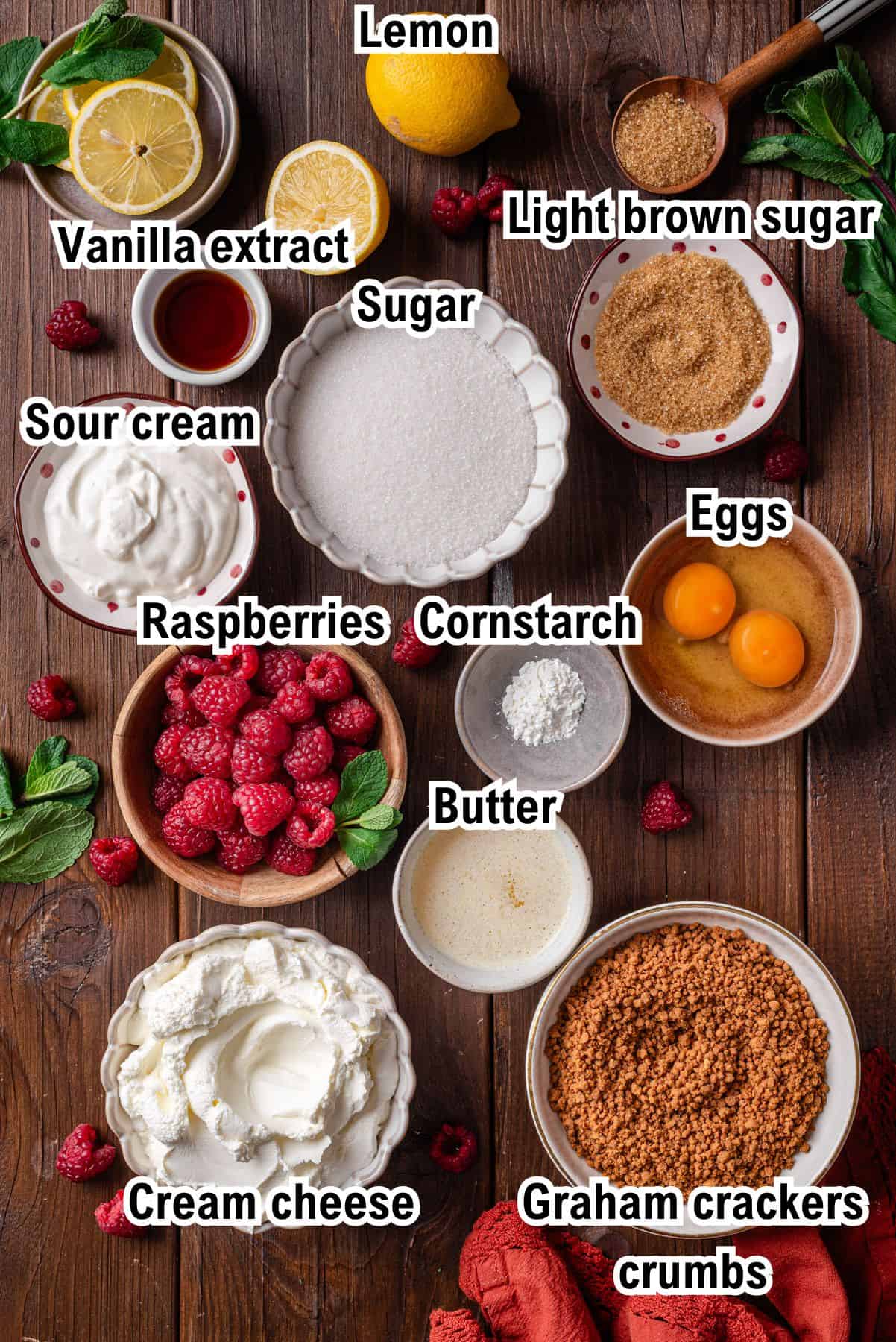 This photo shows the ingredients for Raspberry Cheesecake Bars: Top-down view of labeled cheesecake ingredients in bowls on a wooden table: lemon, vanilla extract, sugar, light brown sugar, sour cream, raspberries, cornstarch, eggs, butter, cream cheese, and graham cracker crumbs.